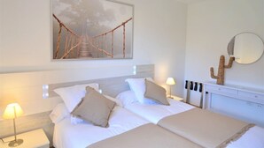 Hotellihuone, jossa on sänky, kaksi yöpöytälamppua, suuri abstrakti maalaus, kaktuskoriste ja pyöreä peili seinällä.
