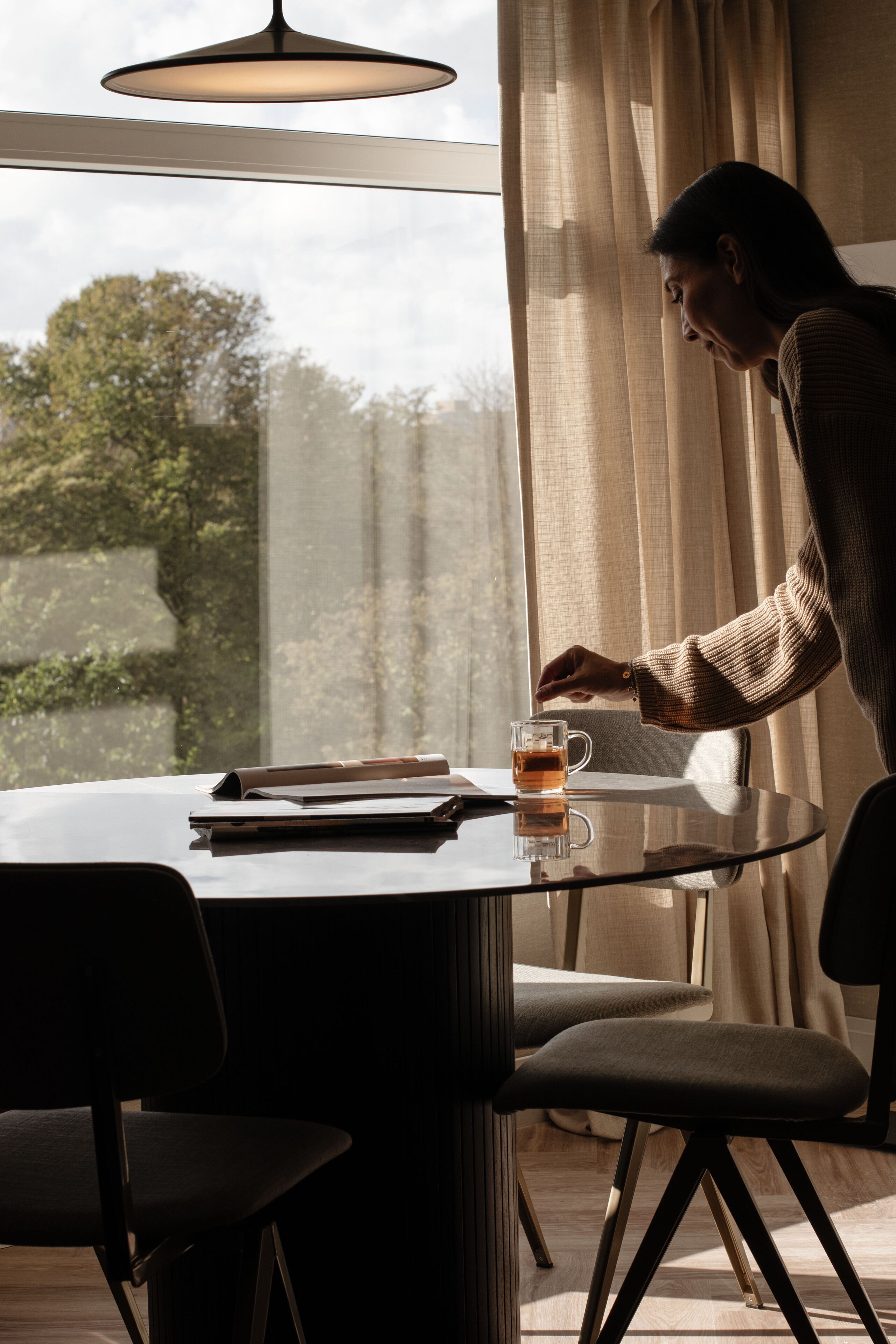 Une pièce moderne avec une table ronde, une tasse de thé et une personne assise près d’une fenêtre avec des rideaux.