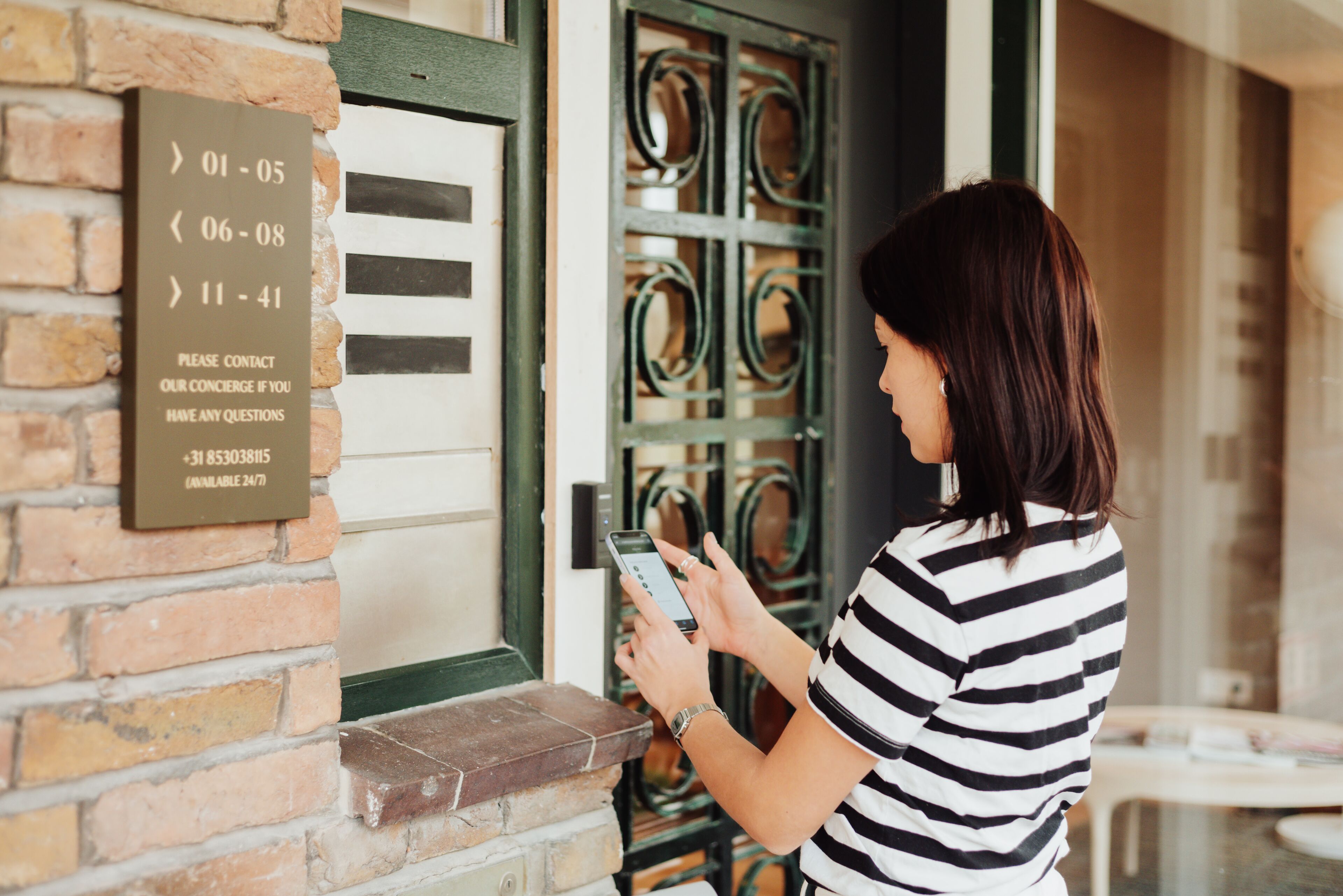 A person standing at a hotel entrance, holding a smartphone, with a sign displaying operating hours and a contact number.