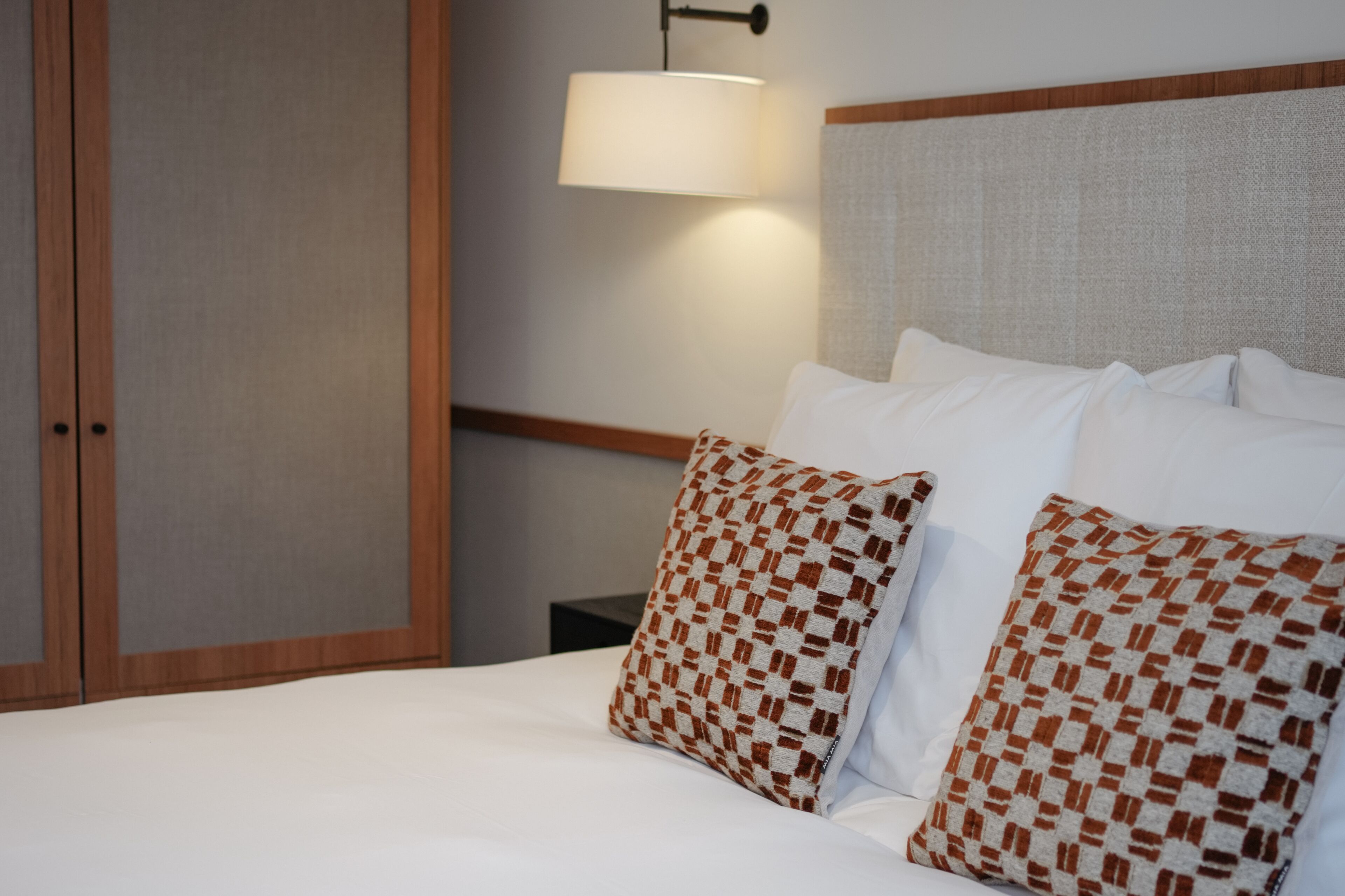 A neatly made bed with white linens and patterned pillows, a wall-mounted lamp, and a wooden headboard.
