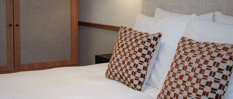 A neatly made bed with white linens and patterned pillows, a wall-mounted lamp, and a wooden headboard.