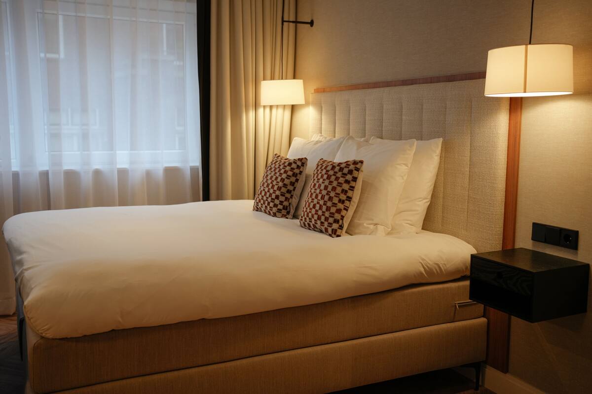 A neatly made bed with white linens and patterned pillows, a bedside table with a lamp, and a window with sheer curtains.