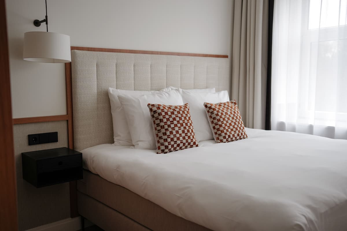 A neatly made bed with white linens and two patterned pillows, a bedside table with a lamp, and a window with curtains.