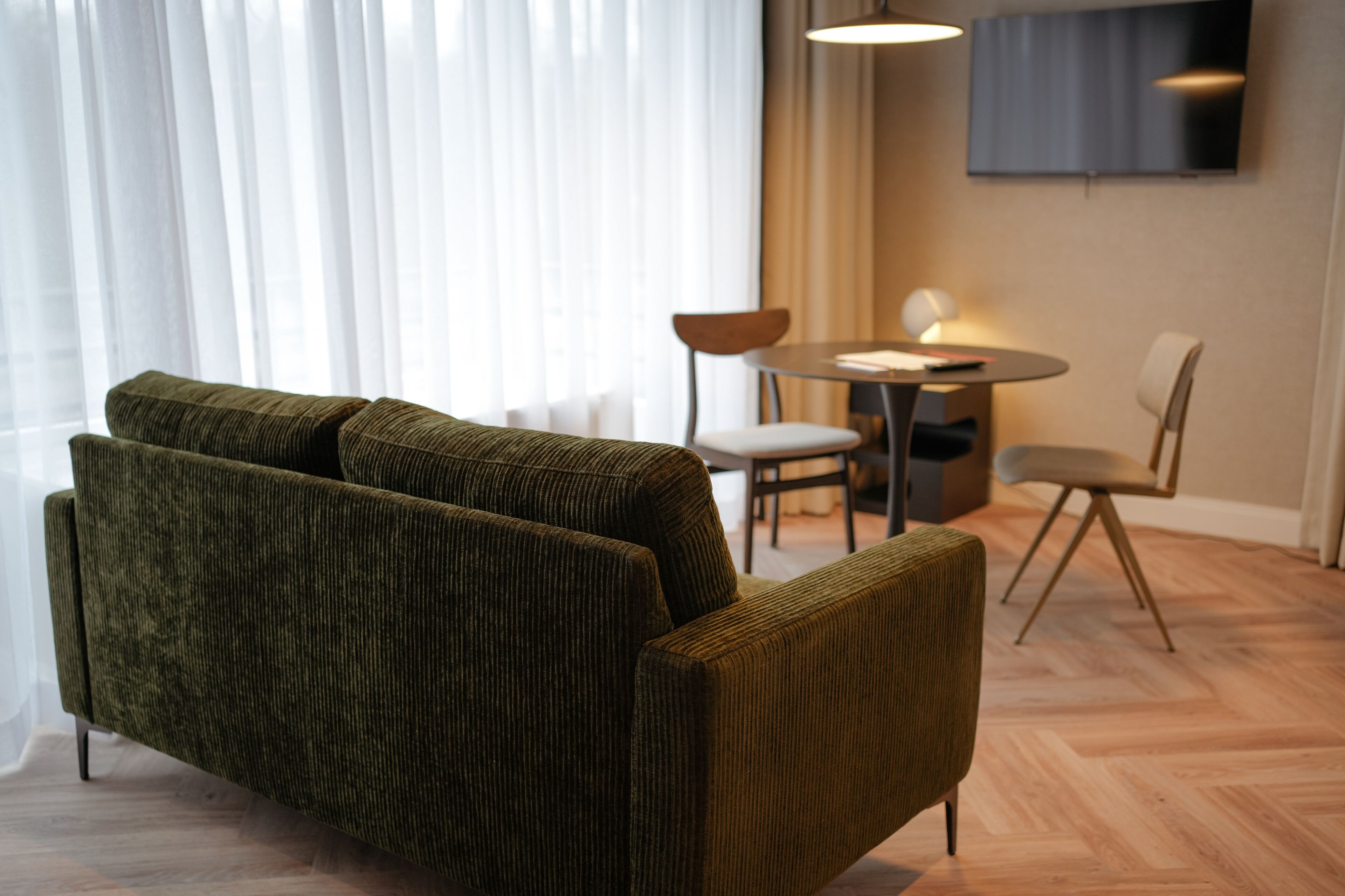 Een moderne woonkamer met een groene bank, een eettafel met stoelen en een aan de muur bevestigde televisie.