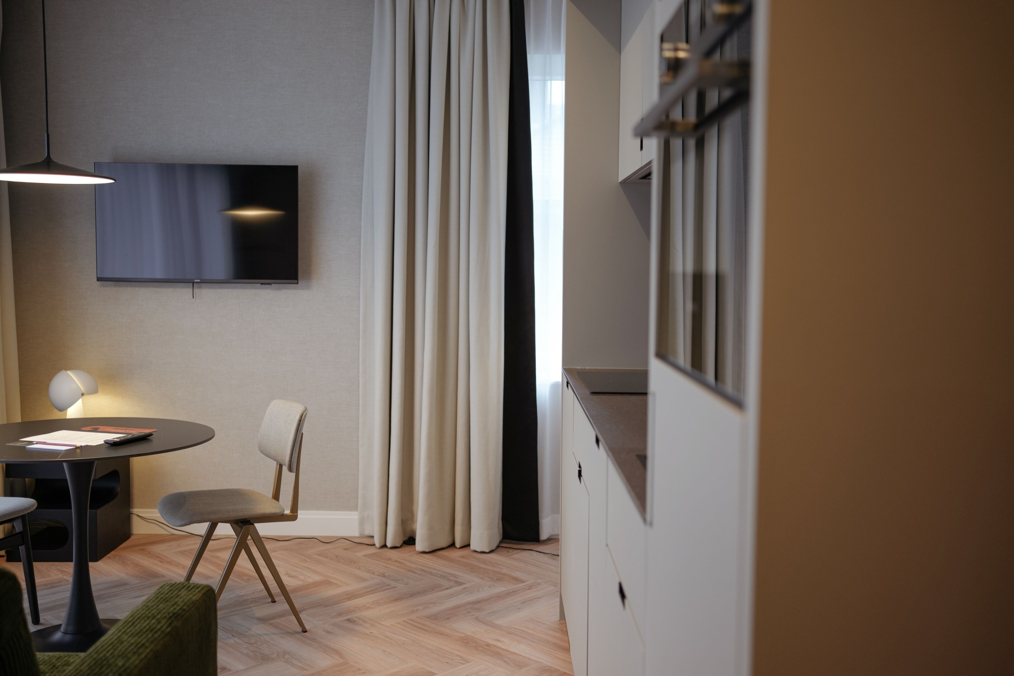 Een moderne kamer met een flatscreen televisie aan de muur, een ronde eettafel met stoelen, een lamp en een groot raam met gordijnen.
