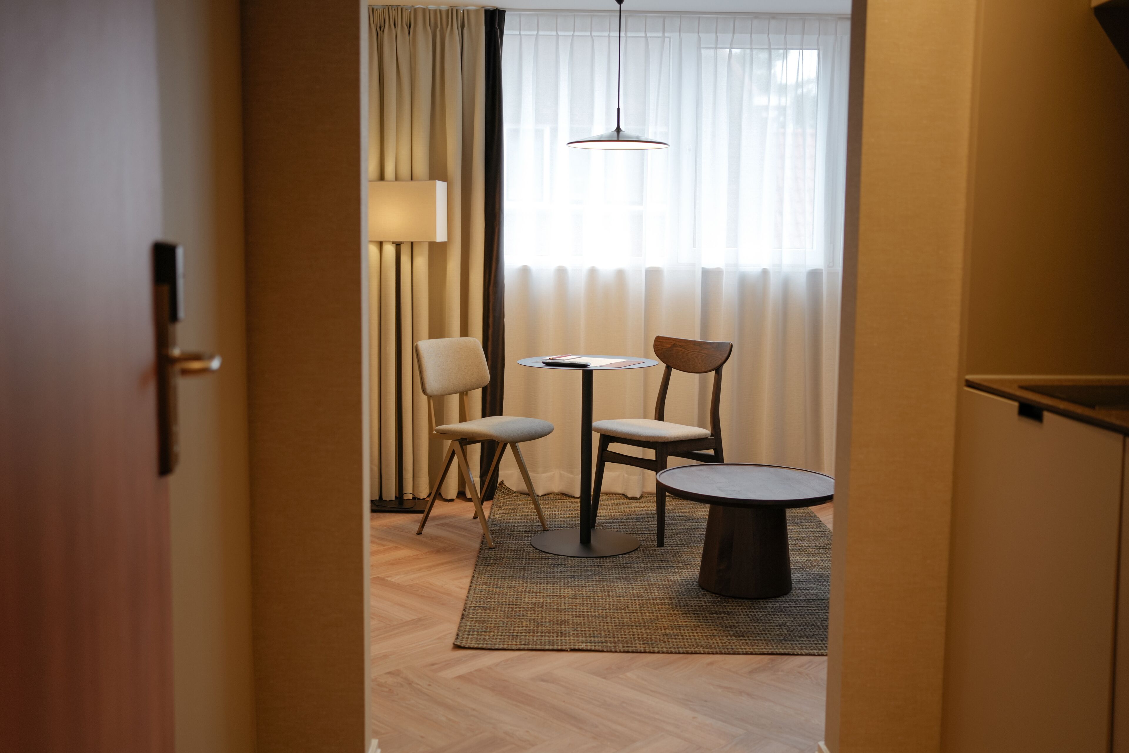 A hallway leading to a dining area with a table and chairs, a lamp, and a view through a window with sheer curtains.