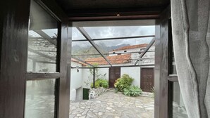 Uitzicht door een raam op een binnenplaats met stenen vloeren, een pergola en traditionele gebouwen met pannendaken.