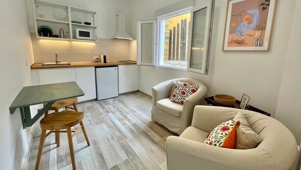 Et kompakt køkken med et spisebord og stole, et lille køleskab og et vindue med udsigt til et hegn. Der er en sofa med puder og et billede i ramme på væggen.