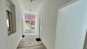 Un pasillo con piso de madera, paredes blancas y una puerta que da a un balcón con flores rosadas.