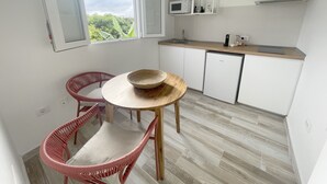 Cocina compacta con armarios blancos, un microondas y un lavamanos. Hay una zona de comedor con una mesa redonda y dos sillas de mimbre.