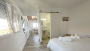 A hotel room with a bed, a small table, and a chair. There is a bathroom with a sink and mirror. The room has a wooden floor and white walls. - Eden Gomera Terrace Apartment oceanviews (Lepe)