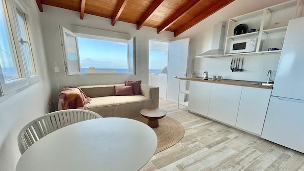 A modern kitchen with white cabinets, a dining area with a round table and chairs, a sofa with cushions, and a view of the sea through large windows. - Eden Gomera Terrace Apartment oceanviews (Lepe)