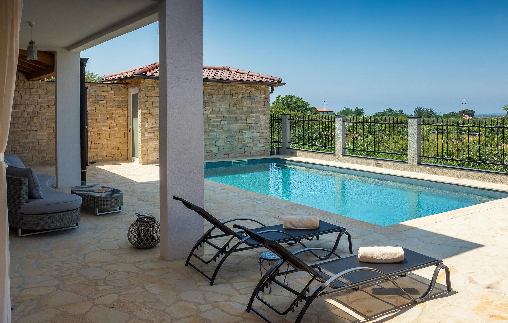A pool area with stone flooring, lounge chairs, and a stone building with a tiled roof.