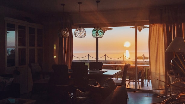 Et værelse med udsigt til en solnedgang over vandet, med et spiseområde med stole og bord, samt en siddeplads med sofa og sofabord.
