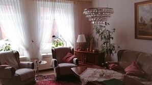 Una sala de estar con alfombra roja estampada, dos sillones, un sofá, una mesa de centro, una araña y un cuadro en la pared.
