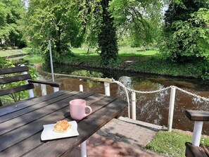 A wooden table with a pink mug and a plate of food, overlooking a river with a stone wall and greenery.