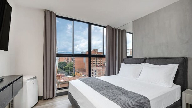 A modern hotel room with a large bed, a desk, and a view of the cityscape through large windows.