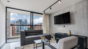 A modern living room with a large window, grey sofa, black coffee table, and a TV mounted on the wall.