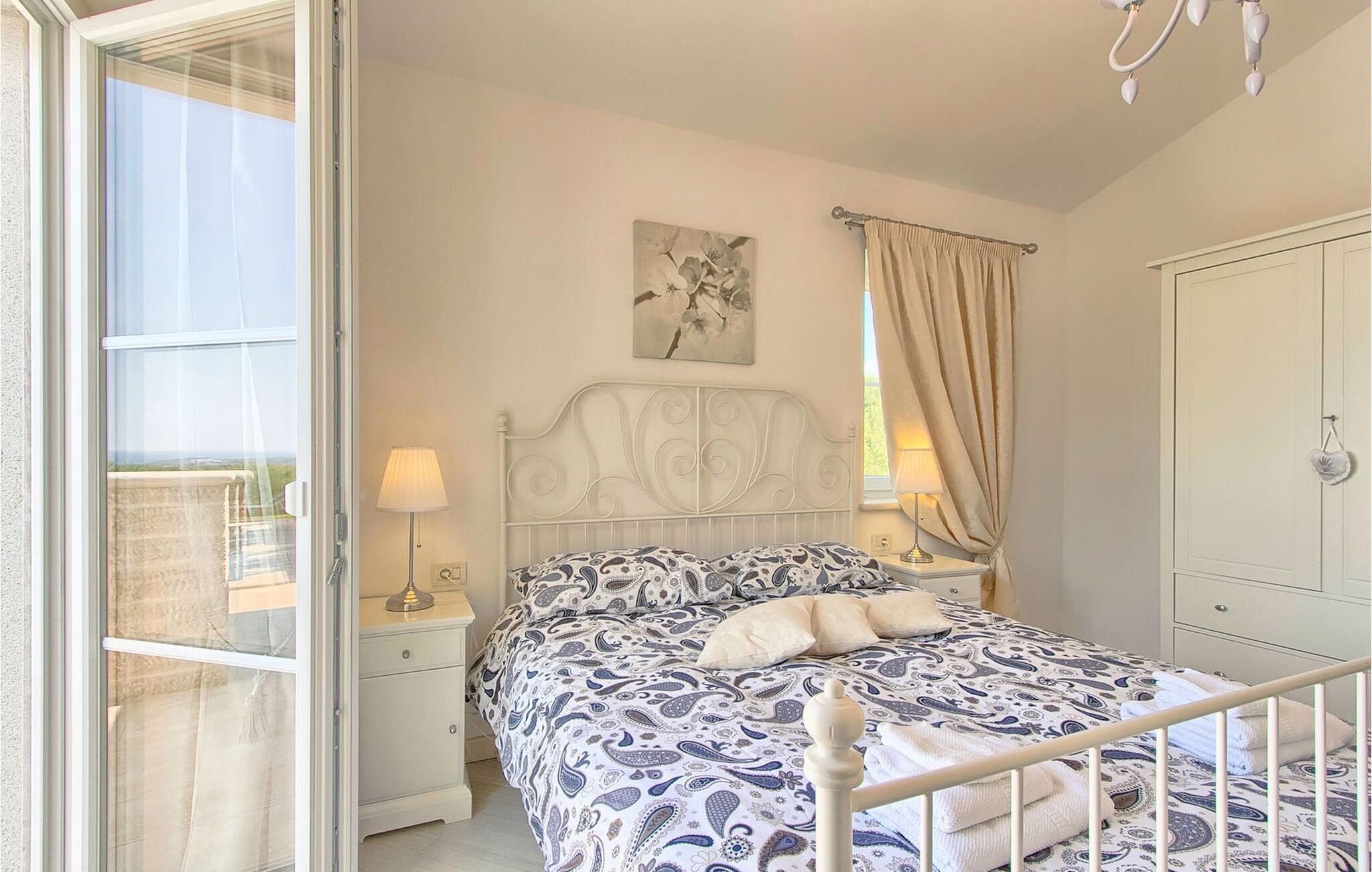 A bedroom with a bed, a nightstand, a lamp, a window with curtains, a painting on the wall, and a wardrobe.