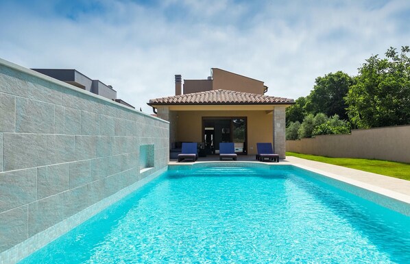Une piscine avec une terrasse carrelée, un patio couvert doté de chaises longues, et une vue sur un immeuble d’habitation en arrière-plan.