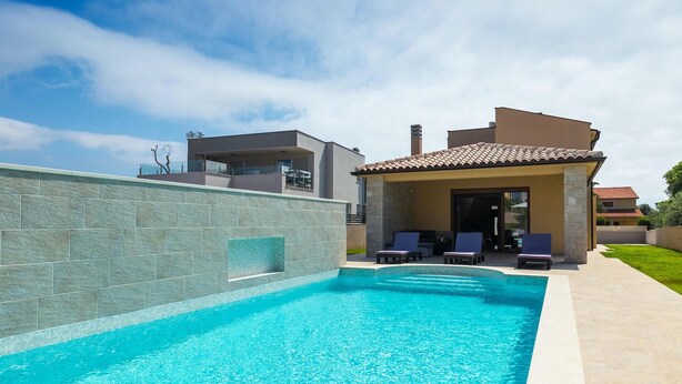 A swimming pool with a stone wall, a covered patio with lounge chairs, and a modern house in the background.
