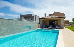 Une piscine bordée d’un mur en pierre, un patio couvert avec des chaises longues, et une maison moderne en arrière-plan.
