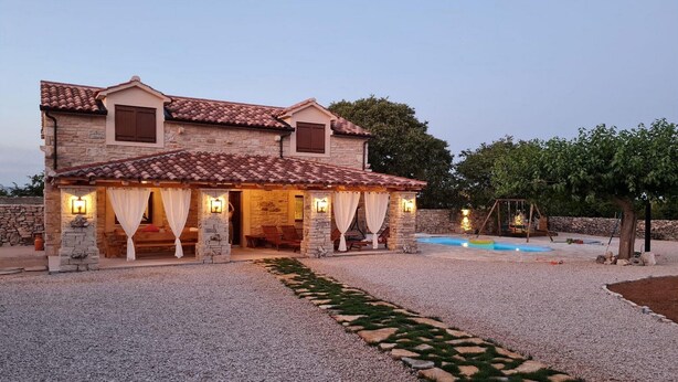 Une maison en pierre à deux étages, avec un toit en tuiles, un patio couvert avec des sièges et une piscine.