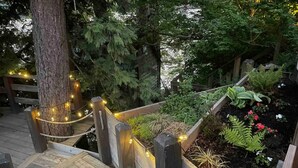 A wooden staircase with string lights, surrounded by lush greenery and a flower bed.