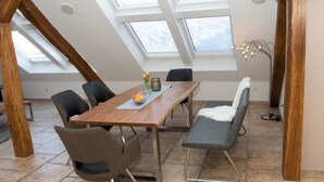 Un espace repas avec une table en bois, des chaises et un banc. Des lucarnes se trouvent au-dessus de la table, et une lampe sur pied est visible.