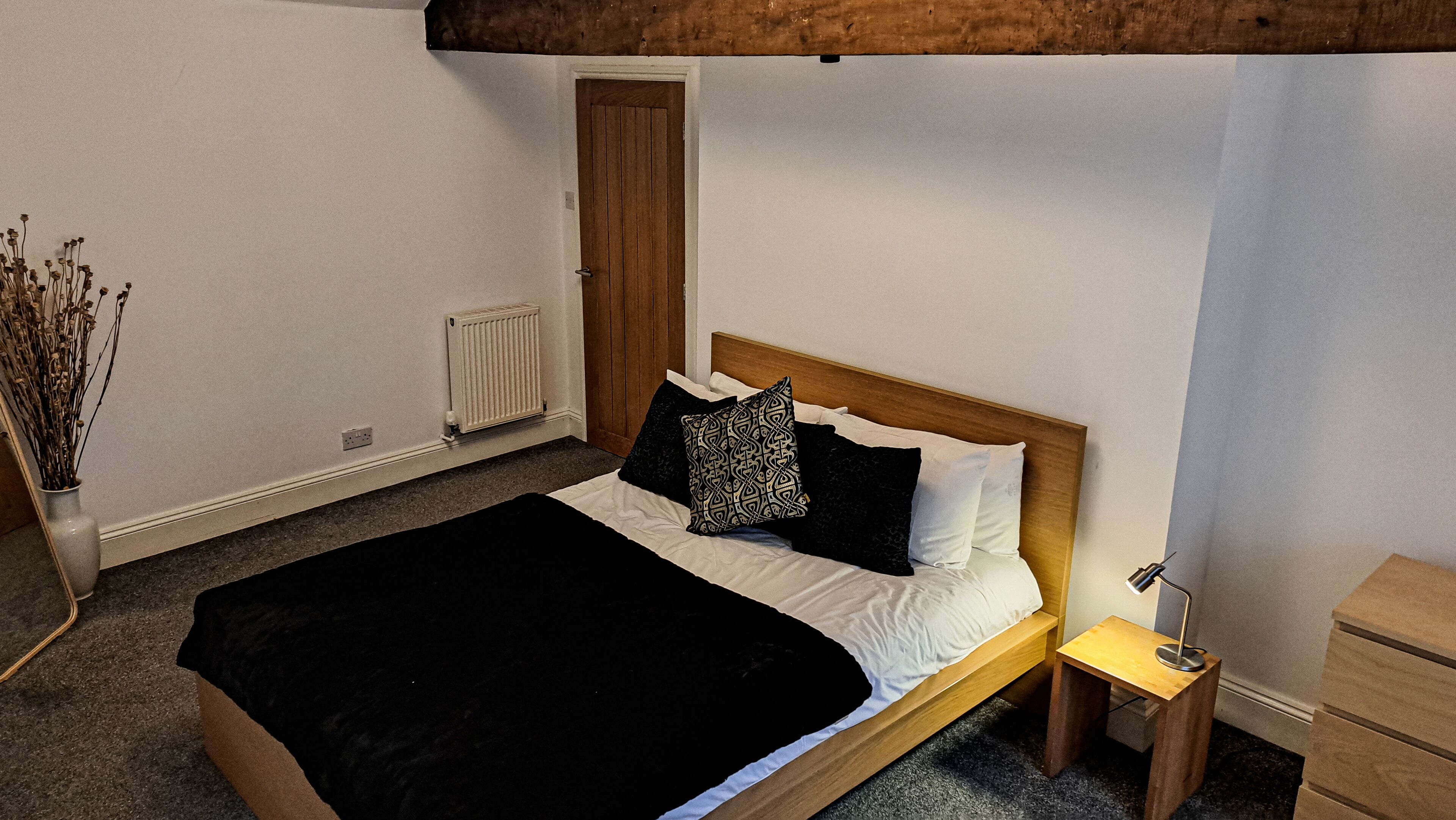 A bedroom with a bed, a nightstand, a wooden headboard, a radiator, a door, and a vase with dried flowers.