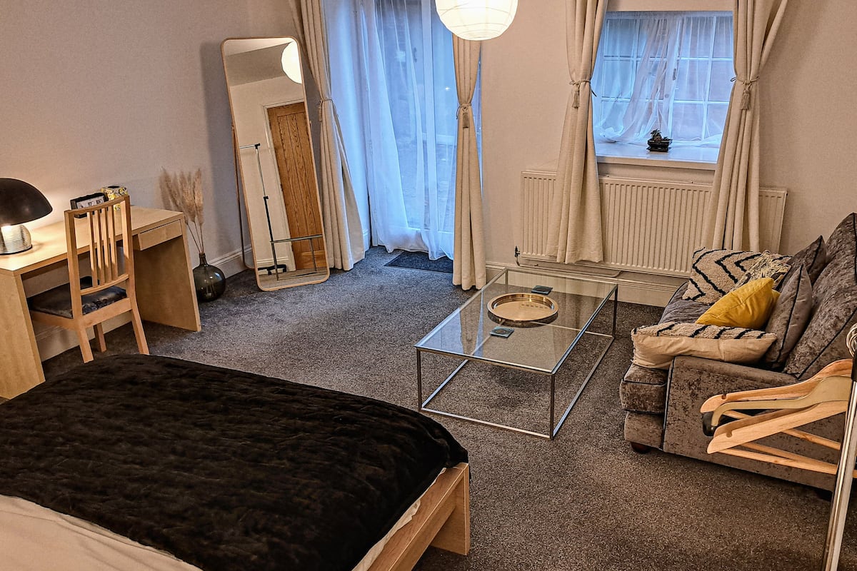 Ett kompakt hotellrum med en säng, en soffa, ett soffbord i glas, ett skrivbord och en stol.