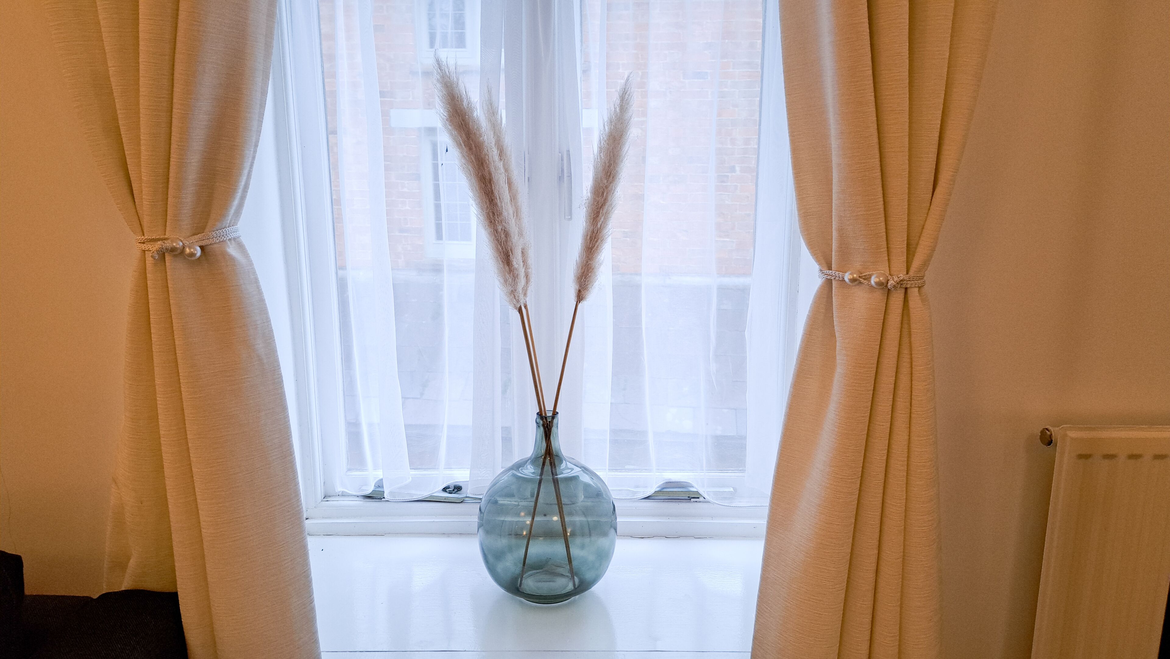 A room with a window, a vase with pampas grass, and curtains.