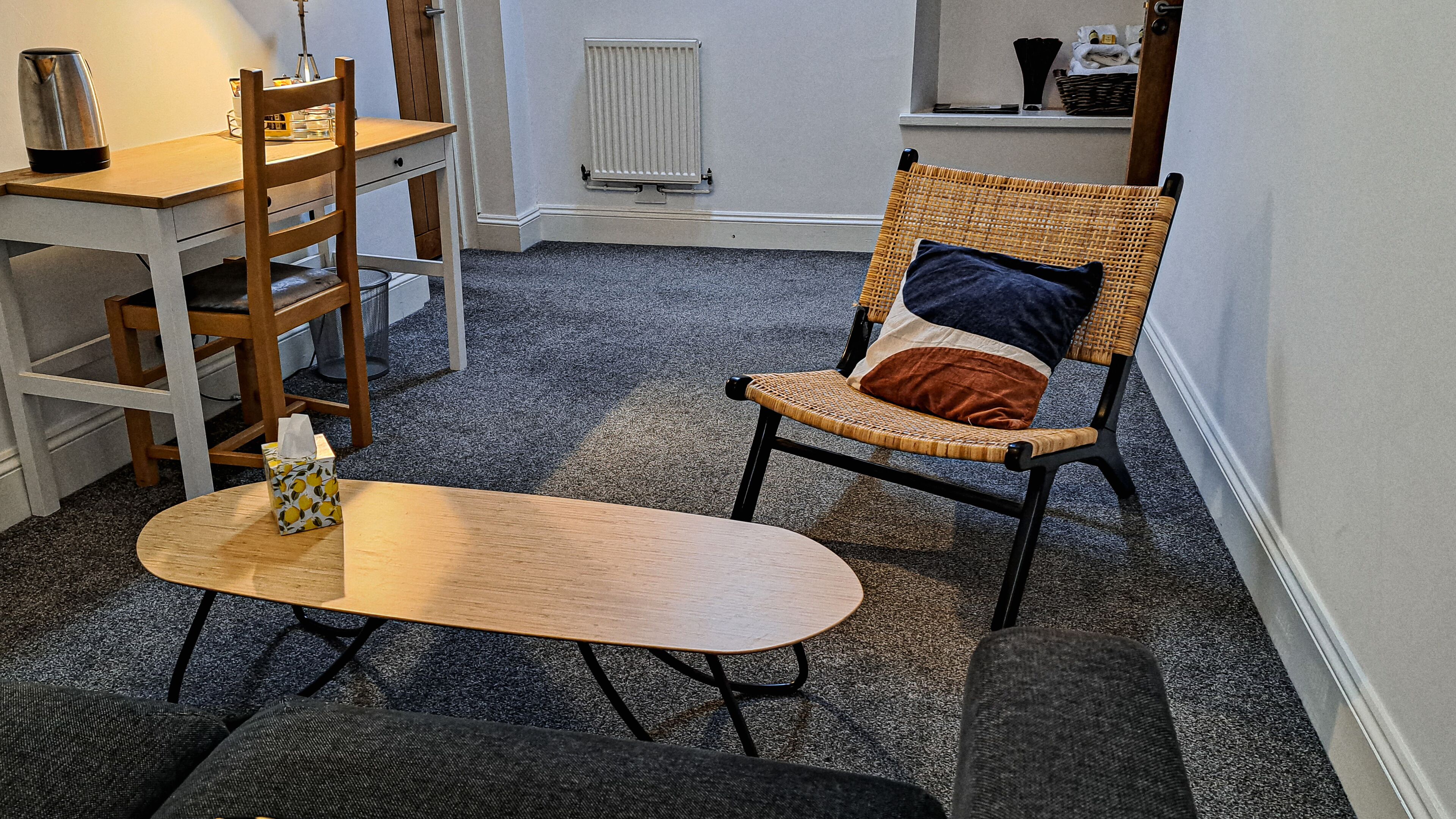 A cozy room with a wooden table, a wicker chair with cushions, a radiator, and a shelf with towels.