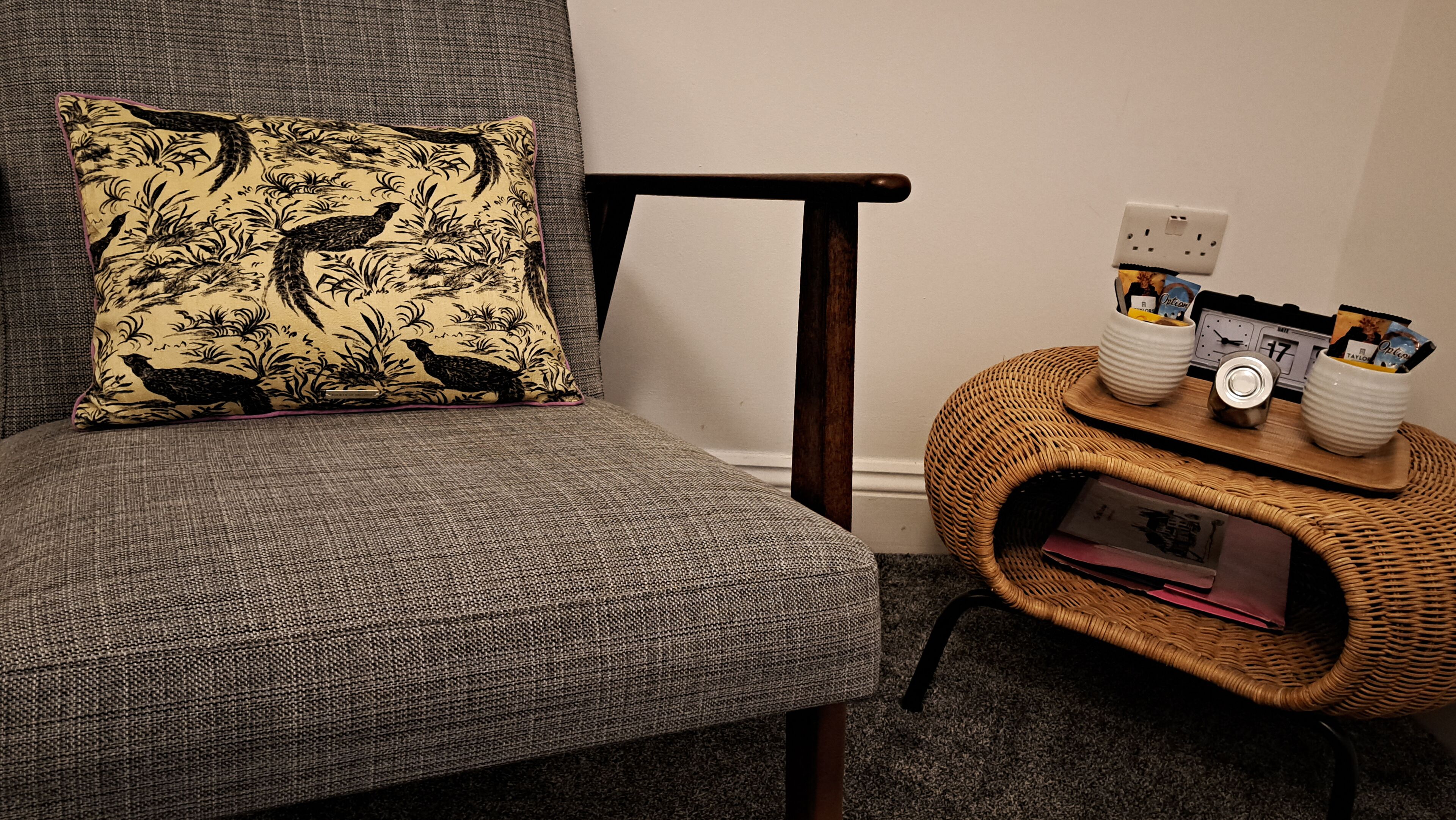 A grey upholstered chair with a floral pillow, a wicker side table with a clock and a small basket, and a power outlet on the wall.