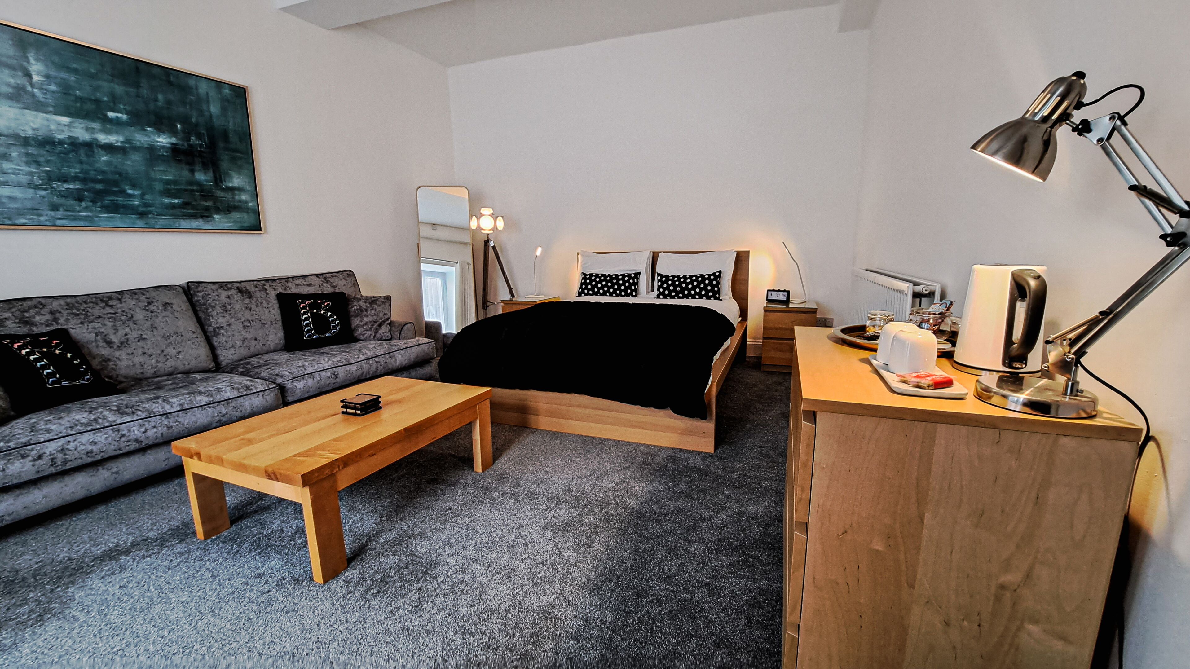 A modern, compact room with a grey sofa, a wooden bed, a small wooden table, a desk with a lamp, and a large abstract painting on the wall.