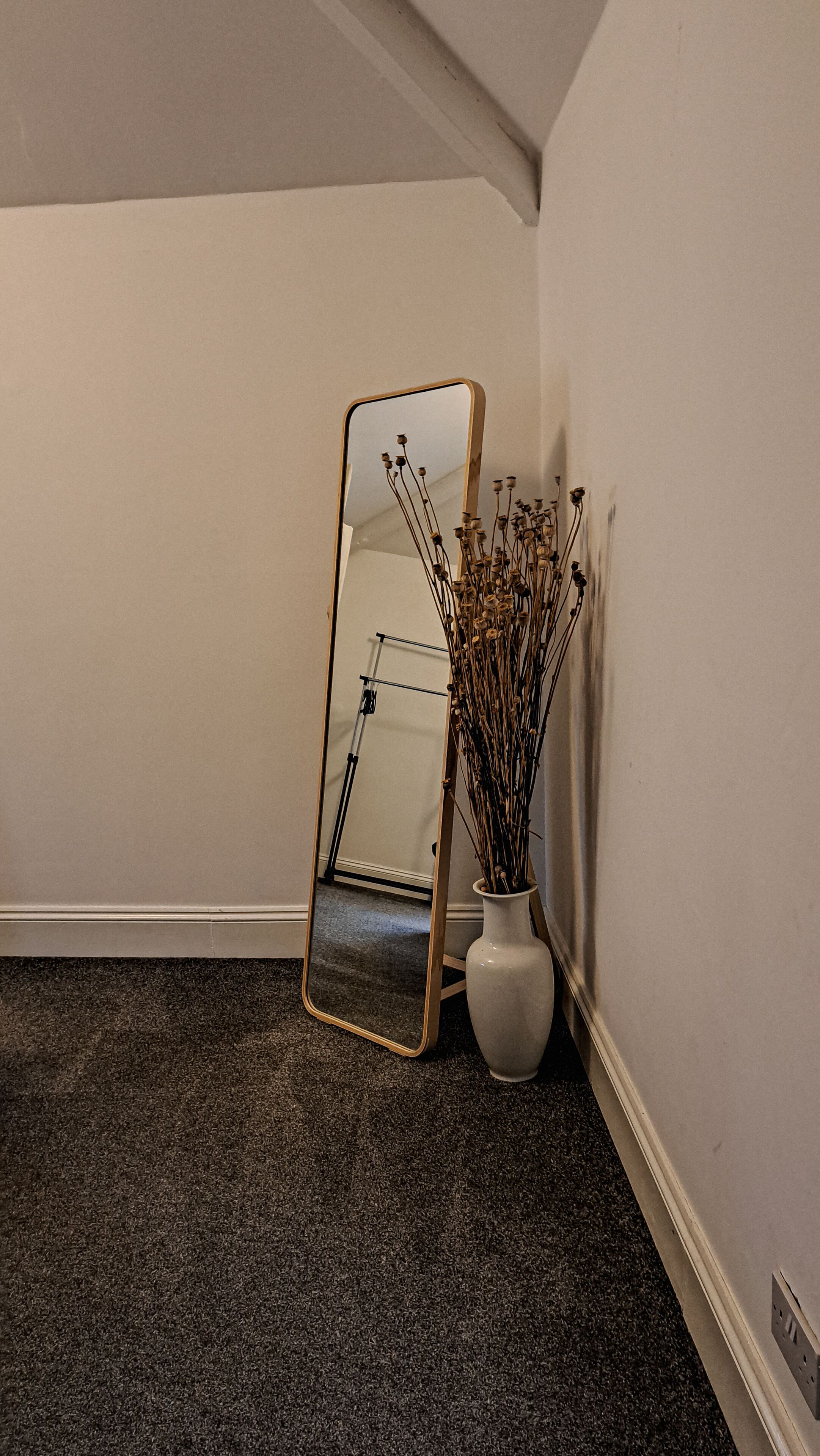 A corner room with a large mirror, a white vase with dried flowers, and a dark carpet.