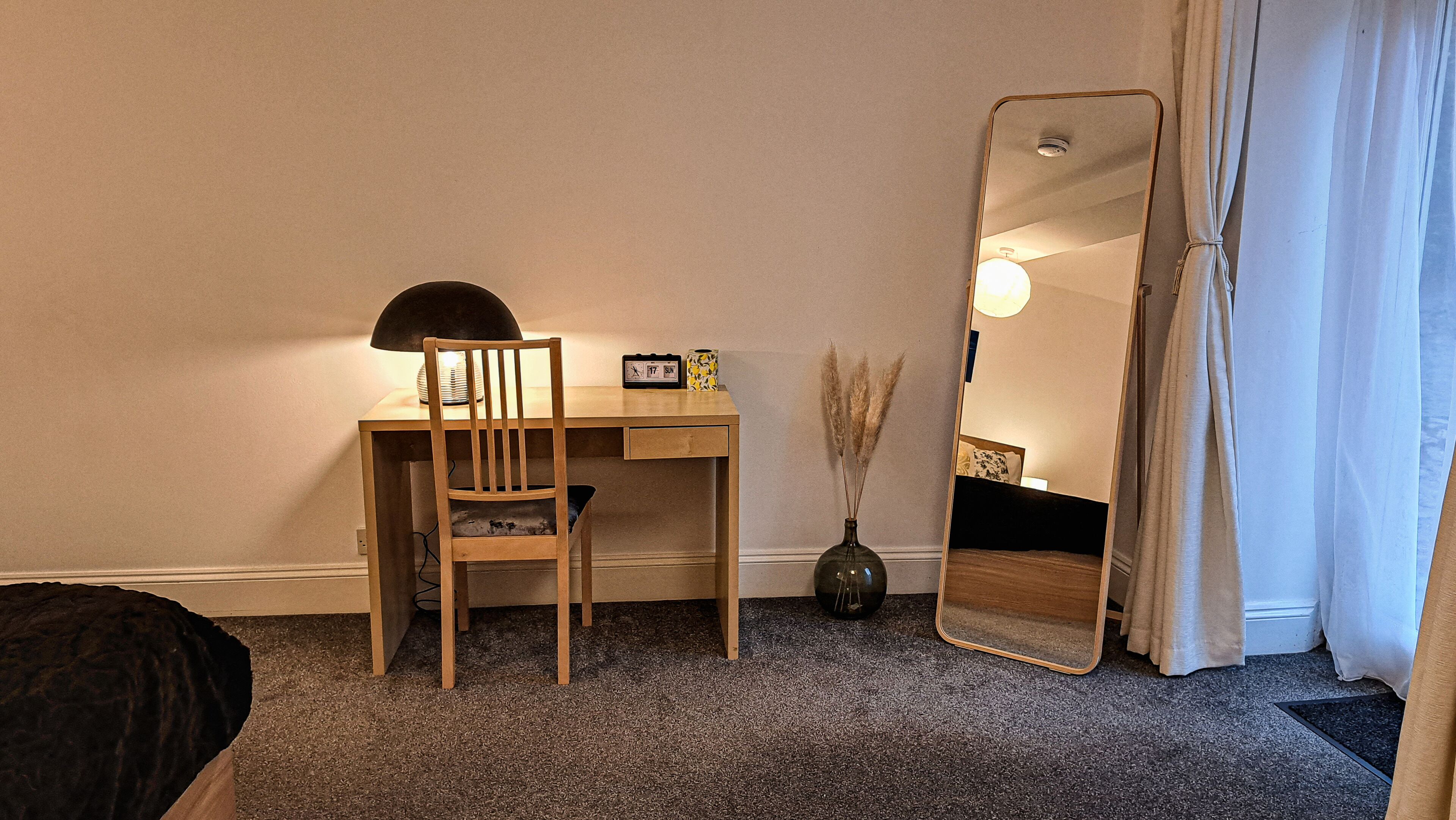 Una habitación con un escritorio de madera, una silla, un espejo, un reloj y un florero con plumas de pampa.