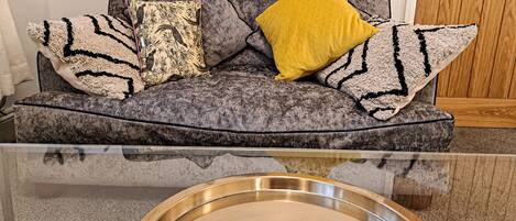 A grey sofa with patterned cushions and a yellow pillow, a glass coffee table with a metal tray, and a peacock feather decoration.