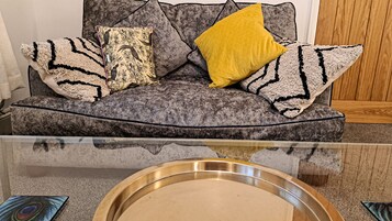 Un sofá gris con cojines estampados y una almohada amarilla, una mesa de centro de vidrio con una bandeja metálica y una decoración con pluma de pavo real.