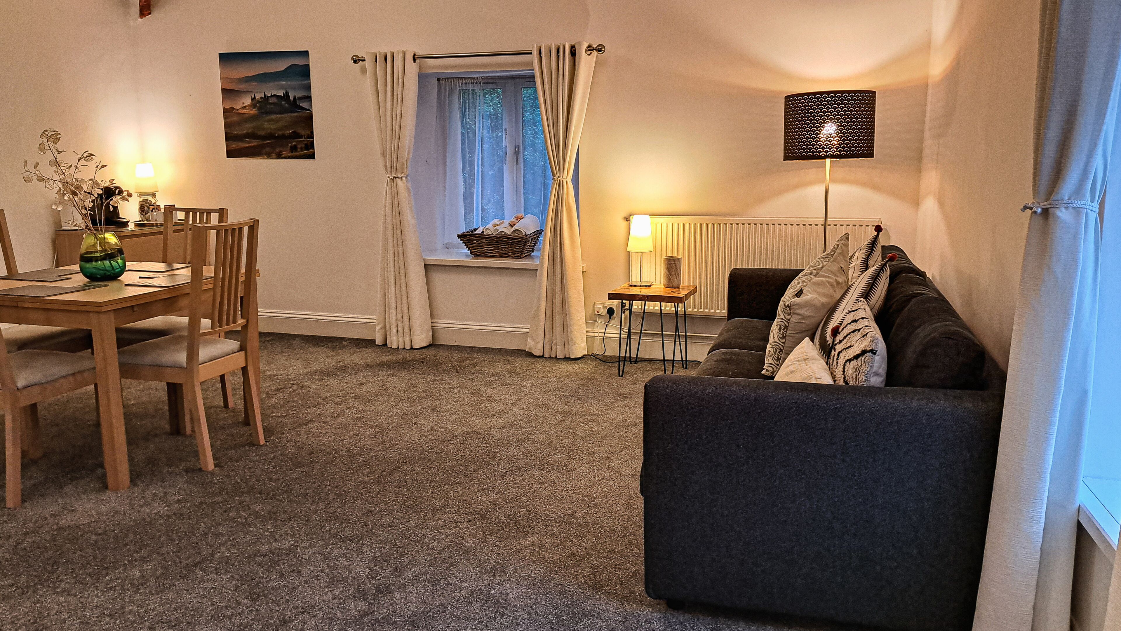 A cozy living room with a sofa, a dining table with chairs, a lamp, and a window with curtains.