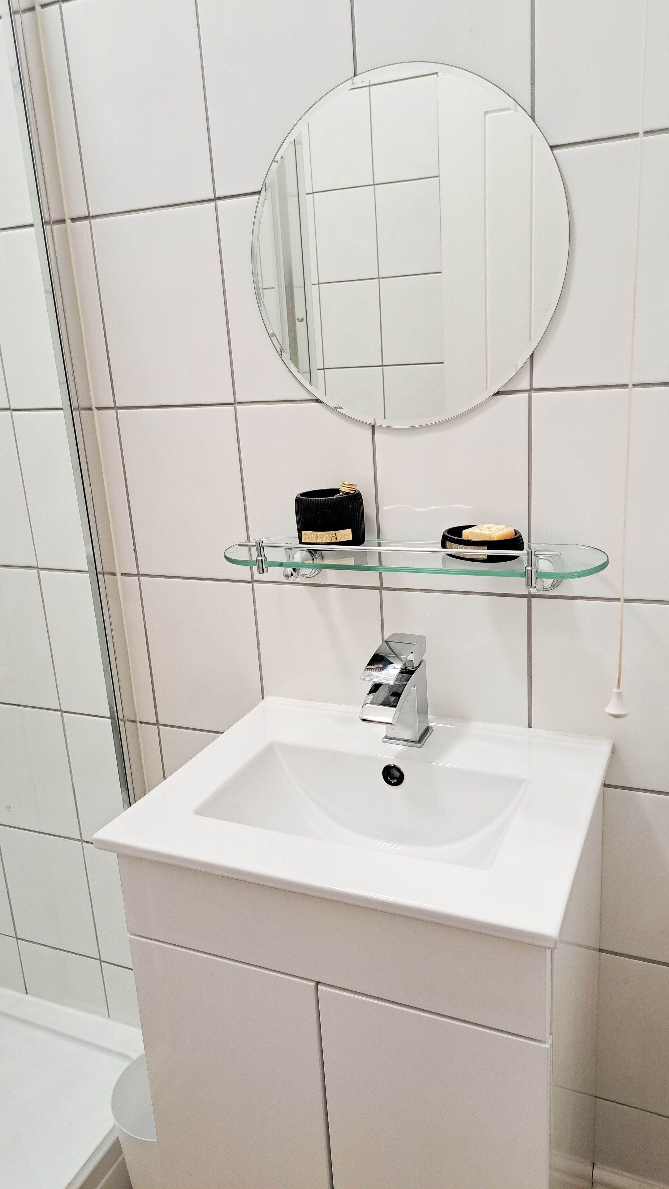 A bathroom with a white sink, a round mirror, and a glass shelf with toiletries.