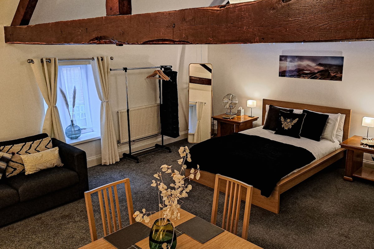 Ett sovrum med en säng, sängbord, ett litet bord, en byrå, ett fönster med gardiner, ett träpanelat tak och ett litet bord med en vas.