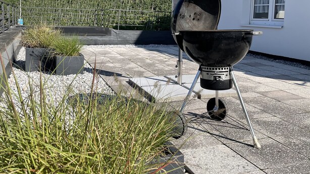 Un barbecue installé sur une terrasse, avec vue sur un vignoble et un bâtiment blanc en arrière-plan.