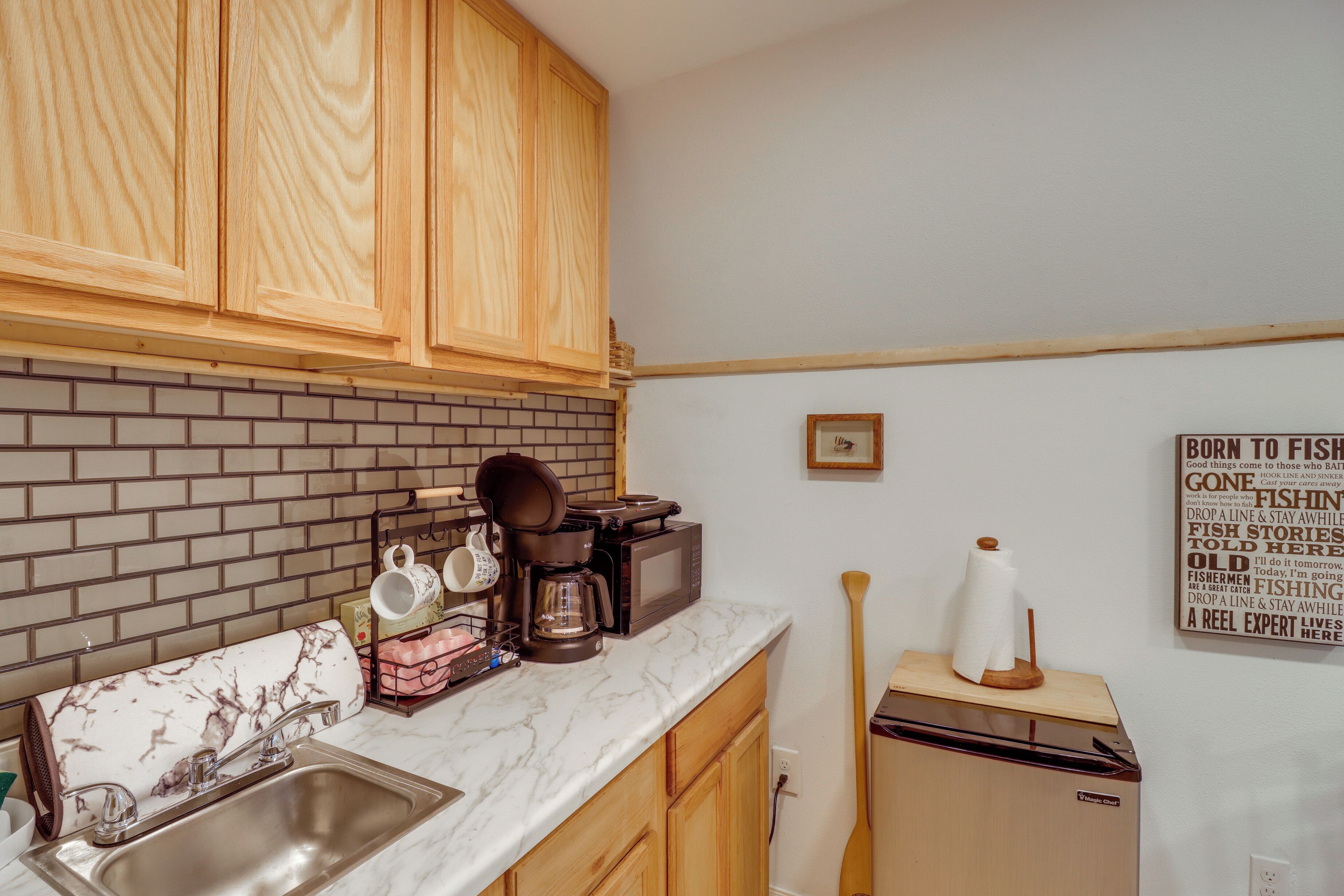 A kitchen with wooden cabinets, a brick backsplash, a marble countertop, a coffee maker, and a sign on the wall.