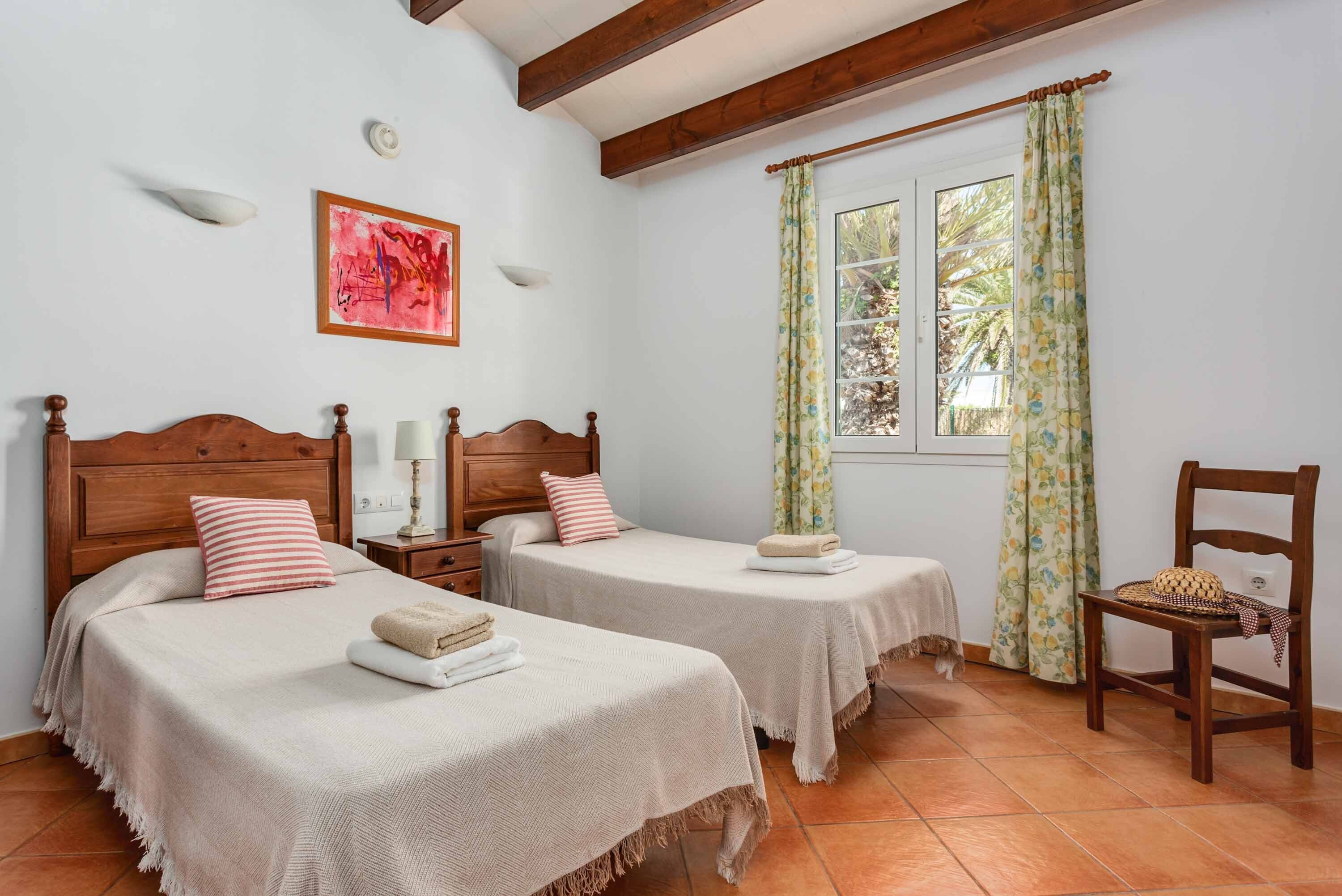 A bedroom with two beds, a window with curtains, a wooden chair, and a wall-mounted light fixture.