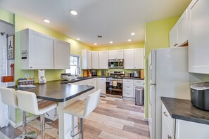 Uma cozinha moderna com armários brancos, uma ilha central com banquetas, eletrodomésticos de aço inoxidável e piso ladrilhado.