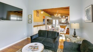 A living room with a dark green sofa, a coffee table with magazines, and a TV on the wall. There is a kitchen in the background with wooden beams on the ceiling. - Inviting Quincy Cottage: Walk to Historic Downtown (Quincy)