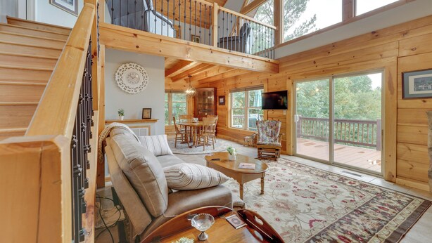 A spacious wooden cabin with a loft bedroom, a cozy living room with a sofa and coffee table, and large windows offering views of the outdoors. - Deck, Grill & Fire Pit: Blue Ridge Mountain Cabin! (Bostic)