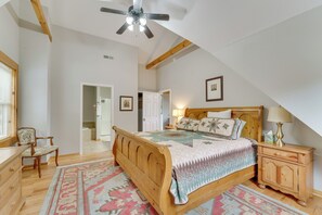 Quarto com cama de madeira, mesa de cabeceira, uma cadeira, ventilador de teto e porta que dá para outro cômodo.