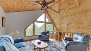A spacious room with a high ceiling, wooden beams, a large window, a blue sofa, a grey leather chair, and a small table. - Deck, Grill & Fire Pit: Blue Ridge Mountain Cabin! (Bostic)